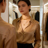 A woman wearing a champagne gold satin button-down shirt and layered gold necklaces, seen looking down at her reflection in a boutique mirror.