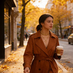 A woman wearing a brown trench coat and gold necklaces walks on a city street with autumn trees and fallen leaves in the background, holding a paper coffee cup.