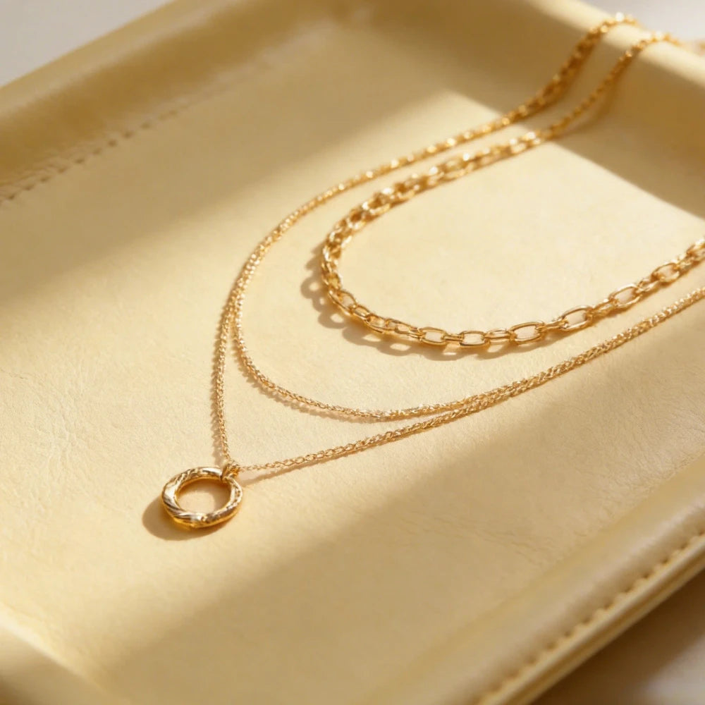 A double-strand gold necklace with a textured circular pendant, displayed on a cream-colored leather tray.