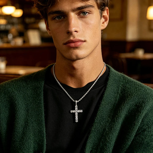 A close-up of a young man wearing a silver cross pendant necklace with clear stones, styled over a black t-shirt and a dark green knit cardigan in a warm, softly blurred indoor setting.