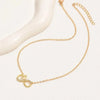 A delicate gold chain necklace featuring a pendant with an infinity symbol and a small heart, accented with tiny clear stones, displayed on a white surface next to a white curved mannequin stand.