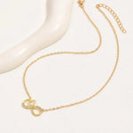 A delicate gold chain necklace featuring a pendant with an infinity symbol and a small heart, accented with tiny clear stones, displayed on a white surface next to a white curved mannequin stand.