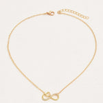 A delicate gold chain necklace featuring a central infinity symbol pendant, one half of which is adorned with small clear stones. The necklace is displayed on a textured off-white background.