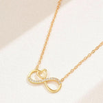 A gold necklace featuring an infinity symbol pendant with a small heart cutout in the center and a row of clear stones along the lower curve, displayed on a textured light beige surface.