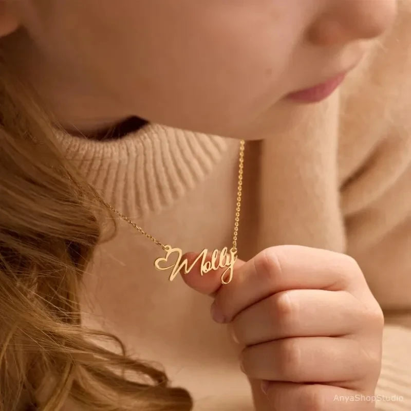 A close-up lifestyle shot of a person wearing a gold name necklace that spells 'Molly' in cursive script, holding one of the letter ends with their fingers.