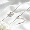 A silver-toned necklace with a teardrop-shaped clear gemstone pendant, displayed on draped white satin fabric with a white flower at the corner.