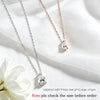 Two heart-shaped crystal pendant necklaces, one silver and one rose gold, displayed on a white fabric background with a white flower in the corner.