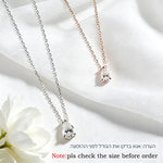 Two heart-shaped crystal pendant necklaces, one silver and one rose gold, displayed on a white fabric background with a white flower in the corner.