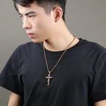 Man wearing a black t-shirt with a gold necklace featuring a cross pendant against a gray background