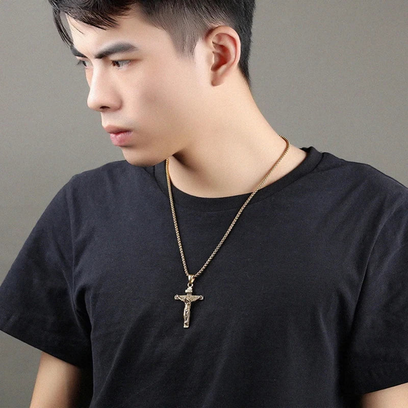 Man wearing a black t-shirt with a gold necklace featuring a cross pendant against a gray background