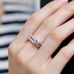 A close-up of a hand wearing a silver-toned double-band ring with a round center stone and small pavé-set stones, shown against a blurred striped background.