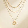 A gold layered necklace set displayed on a cream-colored surface, featuring a twisted oval pendant on a delicate chain, a textured beaded chain, and a classic link chain.