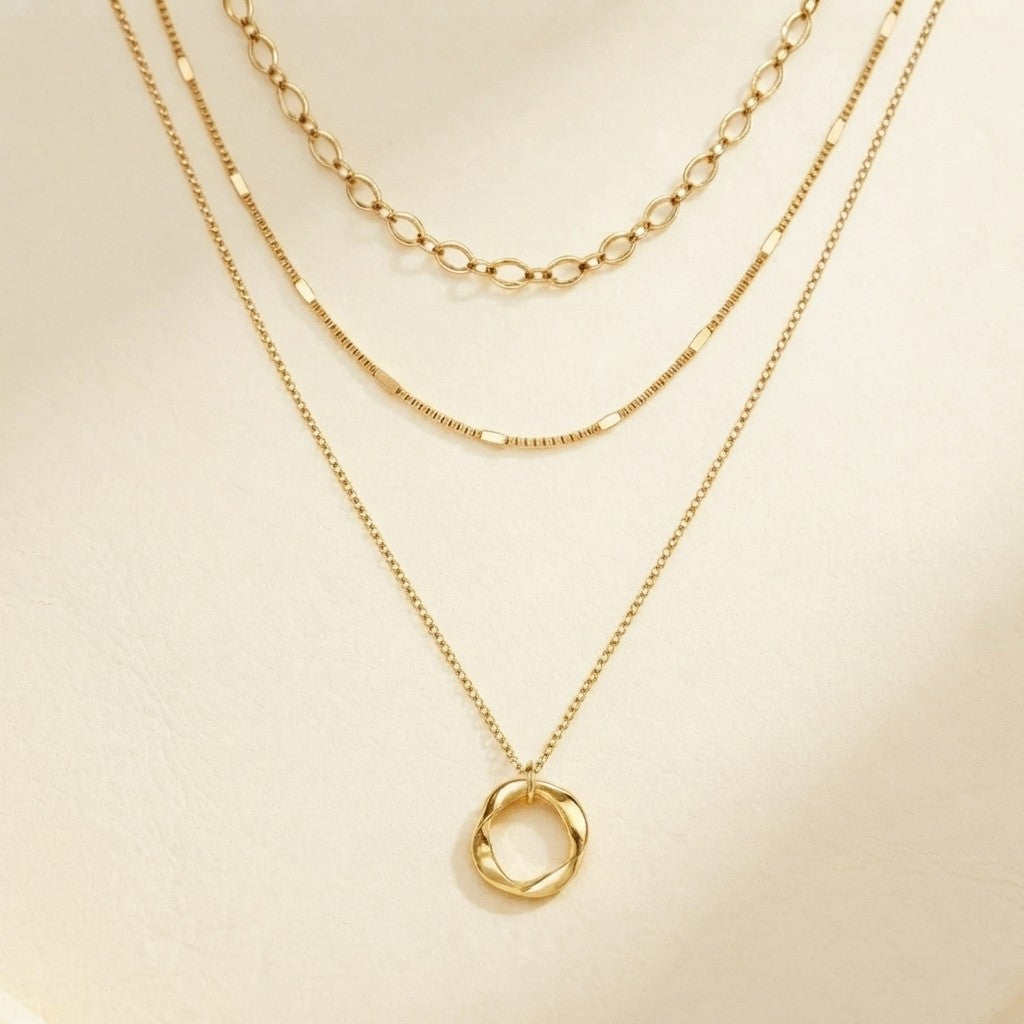 A gold layered necklace set displayed on a cream-colored surface, featuring a twisted oval pendant on a delicate chain, a textured beaded chain, and a classic link chain.