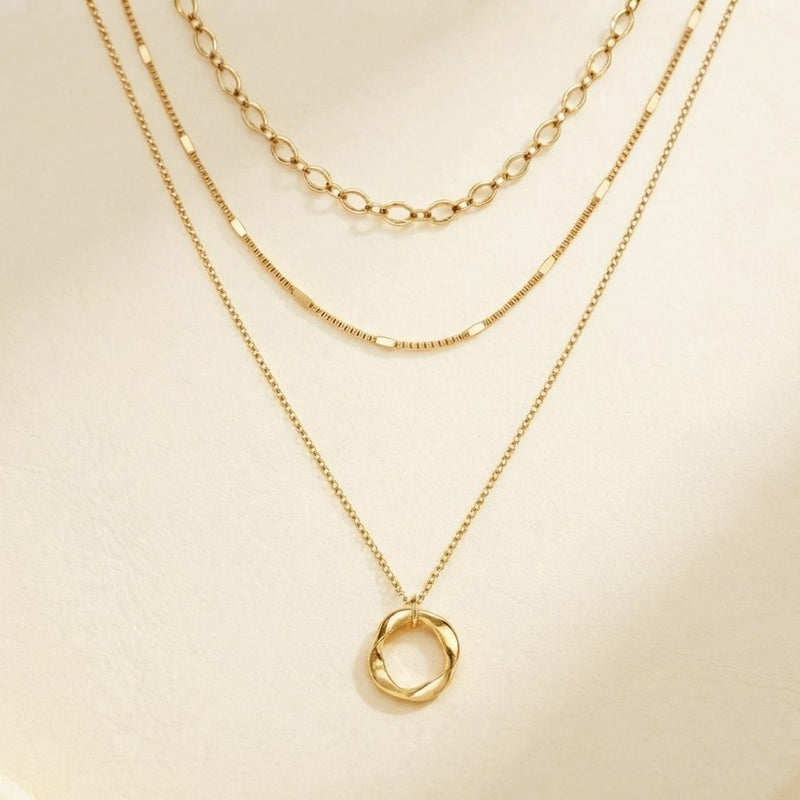 A gold layered necklace set displayed on a cream-colored surface, featuring a twisted oval pendant on a delicate chain, a textured beaded chain, and a classic link chain.