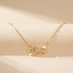 A delicate gold chain necklace featuring a pendant with a gold heart and infinity symbol, accented with small clear crystals, displayed on a soft beige surface.