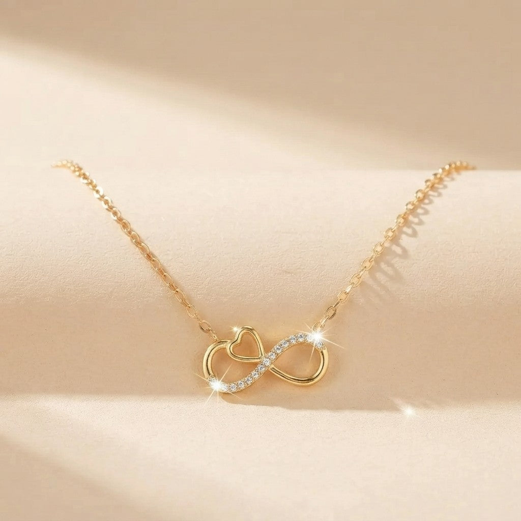 A delicate gold chain necklace featuring a pendant with a gold heart and infinity symbol, accented with small clear crystals, displayed on a soft beige surface.