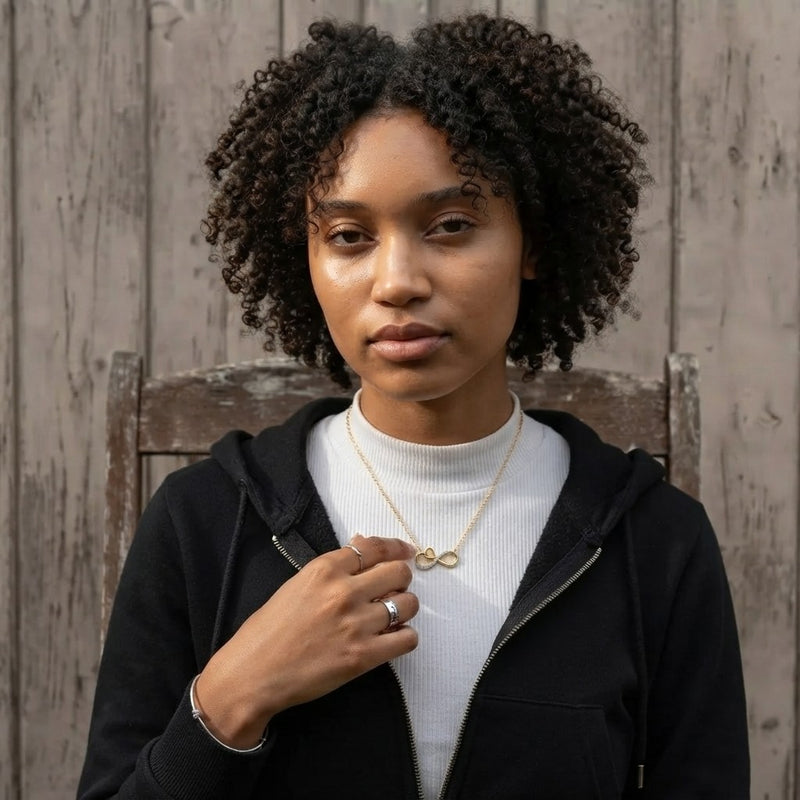 A woman with curly dark hair wearing a black zip-up hoodie over a white ribbed turtleneck, accessorized with a delicate gold infinity necklace and a silver ring, posing in front of a rustic wooden fence.